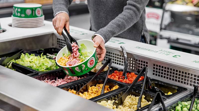 En person plukker salat fra en salatbar med ulike grønnsaker i beholdere.
