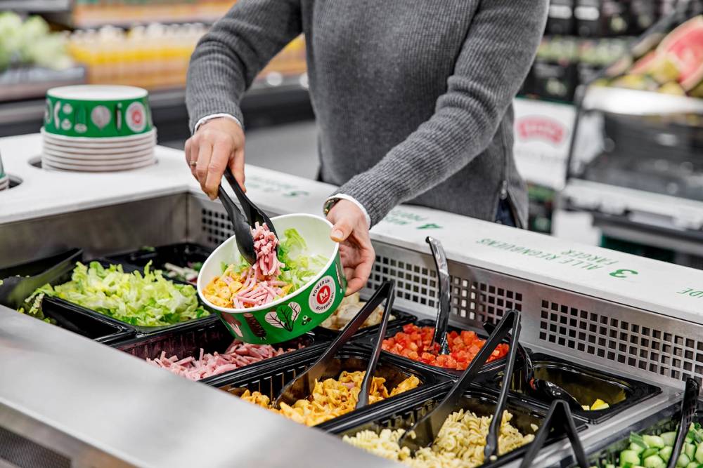 En person plukker salat fra en salatbar med ulike grønnsaker i beholdere.