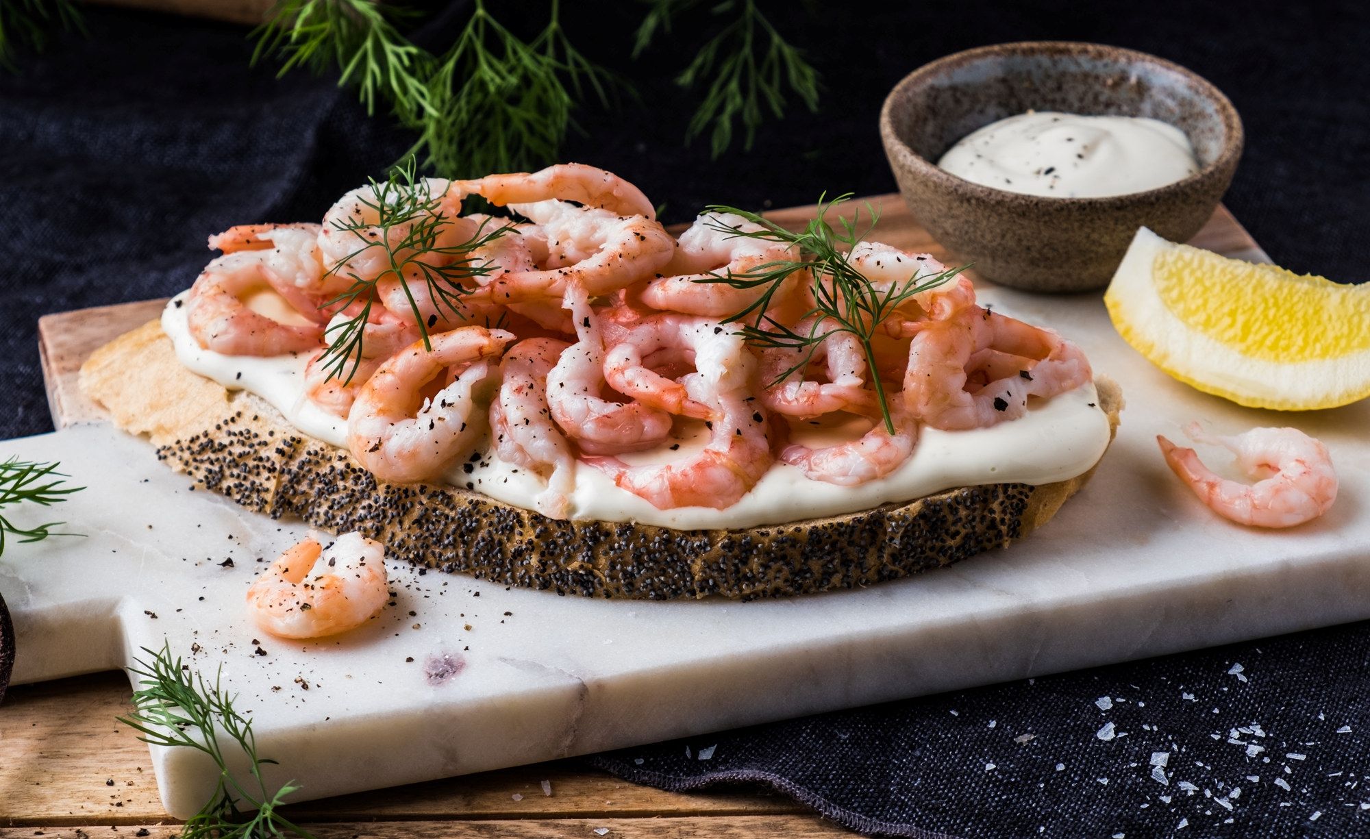 Rekesmorbrod med reker, dill og sitron på en skjærefjøl med en liten skål med saus i bakgrunnen.