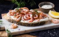 Rekesmorbrod med reker, dill og sitron på en skjærefjøl med en liten skål med saus i bakgrunnen.