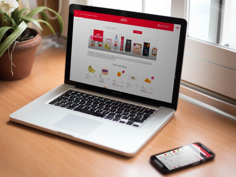 En bærbar datamaskin viser en nettbutikk med ulike produkter på skjermen, plassert på et bord ved et vindu.