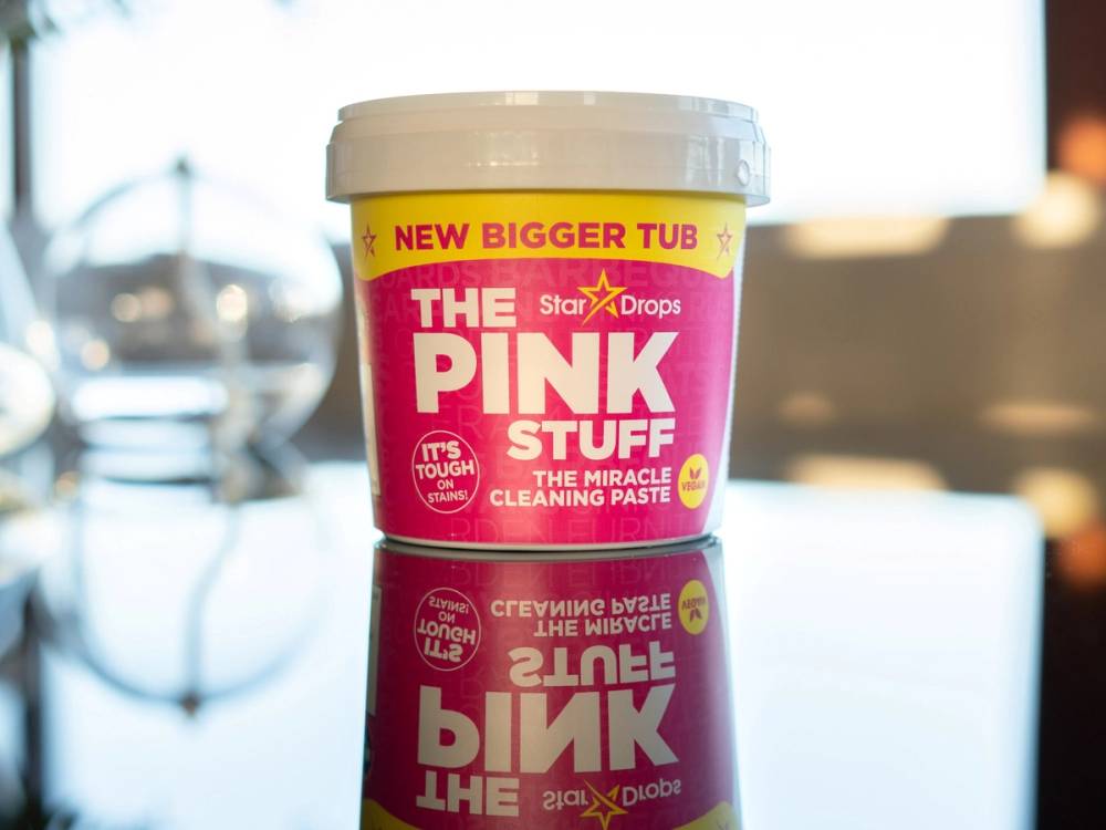 En stor rosa krukke med rengjøringskrem merket 'The Pink Stuff' står på et bord med en dataskjerm i bakgrunnen.