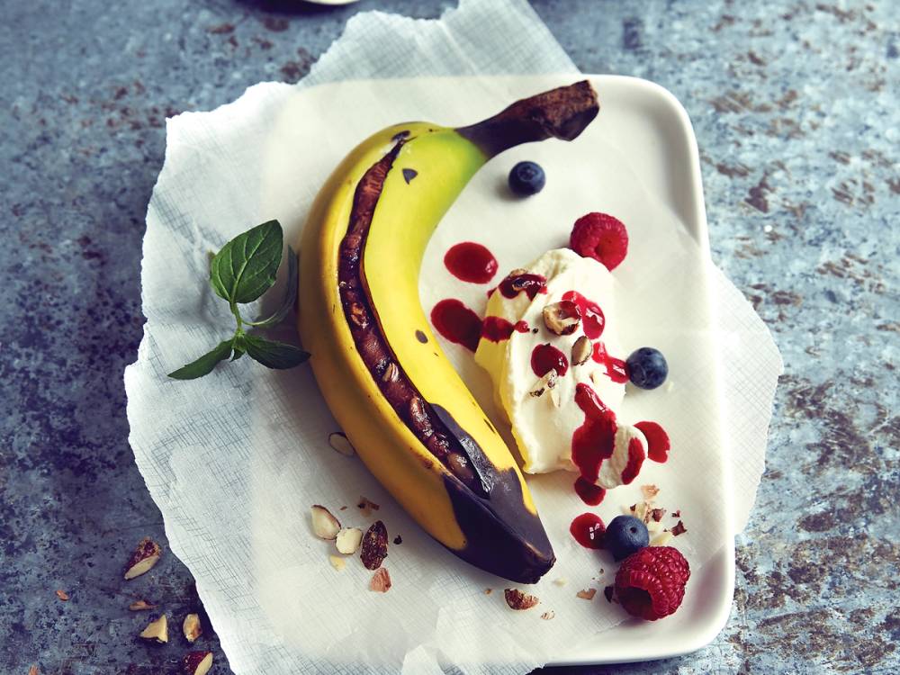 Grillet banansplitt med banan, bær og is på en hvit tallerken, pyntet med mynteblad og nøtter.