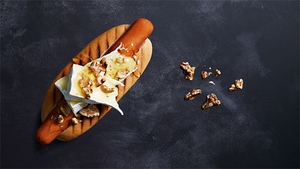 En pølse i brød med ost og nøtter på en treplate, plassert på en mørk bakgrunn.