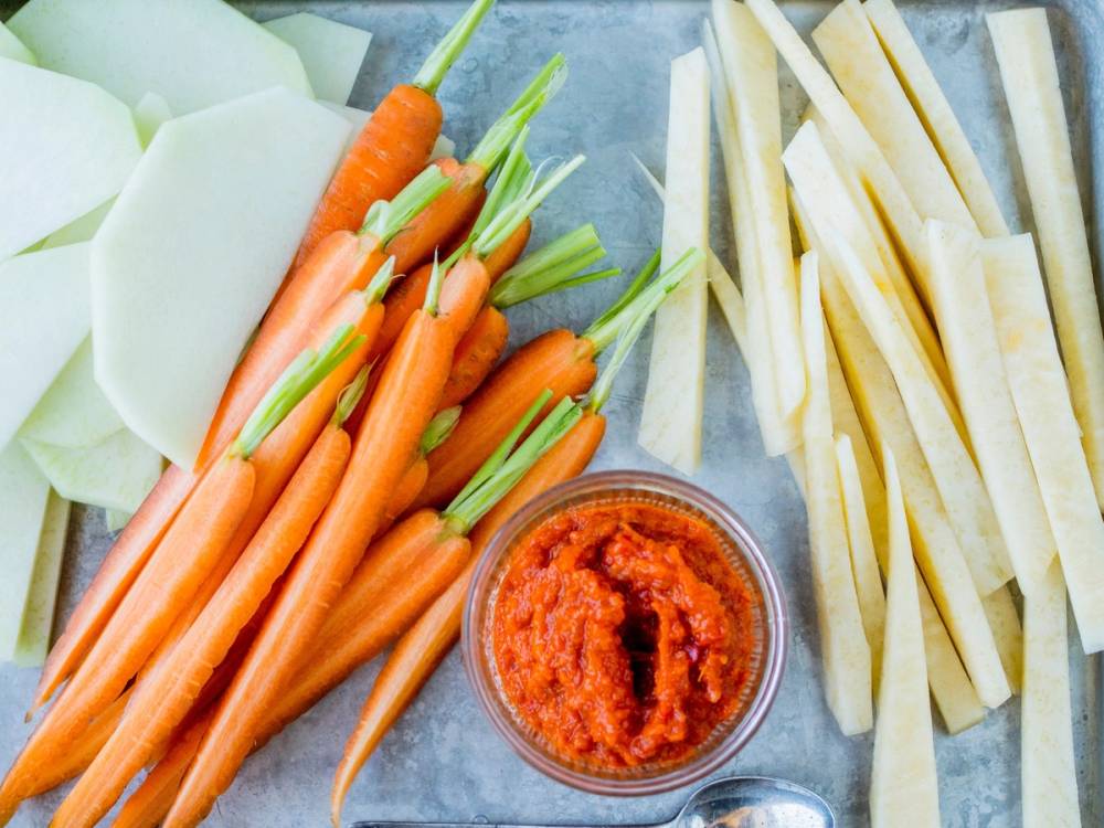 Ferske gulrøtter, selleristenger og en skål med tomatpesto på et skjærebrett.