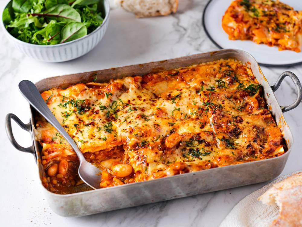 Vegetarisk lasagne i en ildfast form med ost og grønnsaker på toppen, med en skje i formen og en salat ved siden av.