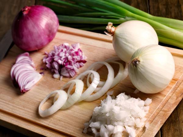 Hvitløk, vårløk og sjalottløk ligger på et skjærebrett sammen med oppkuttet løk og en kniv, klare til bruk i matlaging.