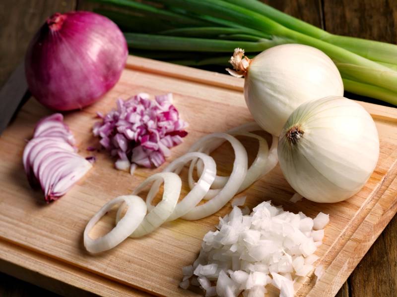 Hvitløk, vårløk og sjalottløk ligger på et skjærebrett sammen med oppkuttet løk og en kniv, klare til bruk i matlaging.
