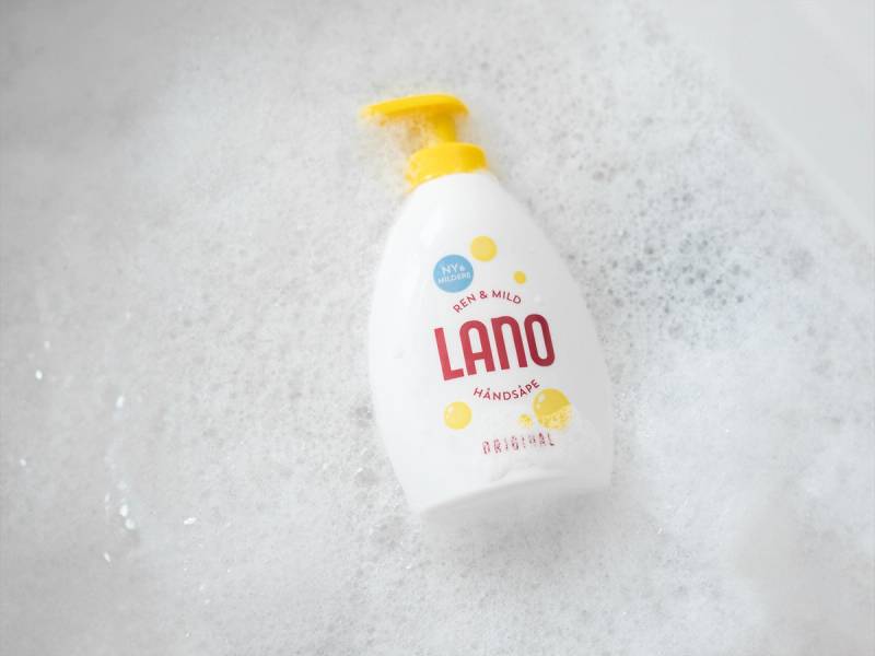En flaske med Lano håndsåpe med gul pumpe på en vask eller benkeplate, omgitt av vann og skum.
