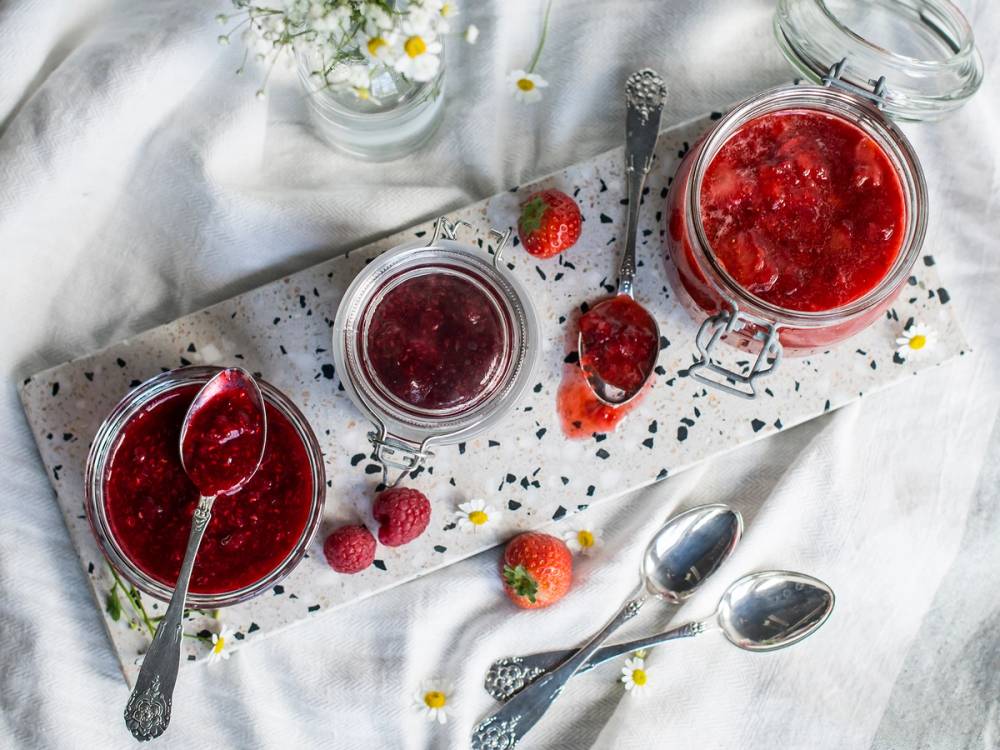 Bilde av tre glass med hjemmelaget syltebærsyltetøy omgitt av friske bær og blomster på et hvitt bord.