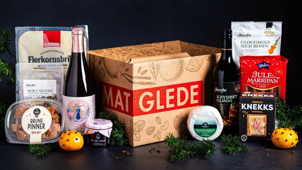 En eske med gaveesker og ulike mat- og drikkevarer, inkludert vin, kjeks og snacks, plassert på et bord.