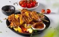 Grillet kjøtt og grønnsaker på spyd med en skål med tomater og en liten bolle med saus ved siden av, på en svart tallerken.