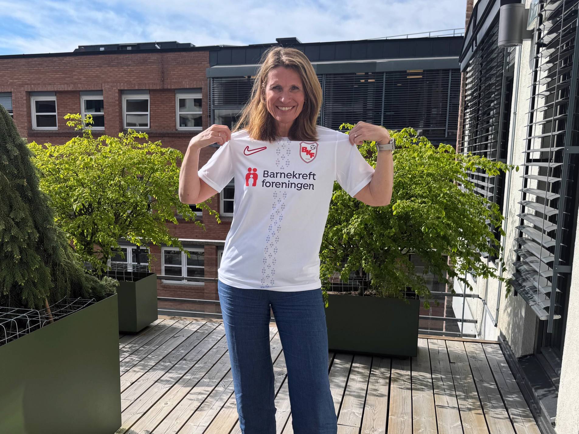 En person står på en balkong med grønne planter og en bygning i bakgrunnen, iført en hvit t-skjorte og blå jeans.