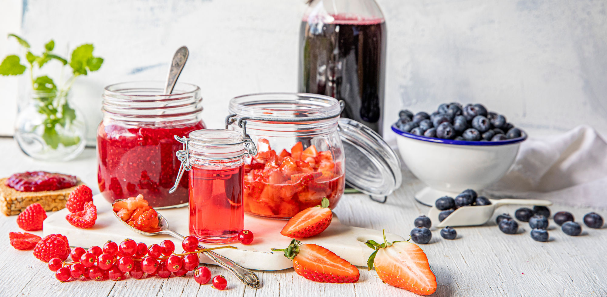 Et utvalg av friske bær, inkludert jordbær, bringebær, blåbær og rips, med en flaske og glass med bærjuice på et kjøkkenbord.