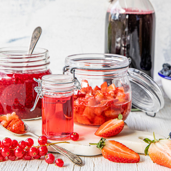 Et utvalg av friske bær, inkludert jordbær, bringebær, blåbær og rips, med en flaske og glass med bærjuice på et kjøkkenbord.