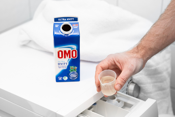 En person holder et lite glass med melk foran en kartong med OMO melk på et hvitt bord.
