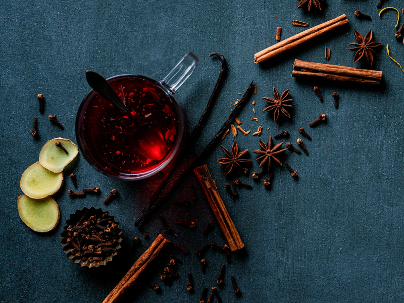 Kopp med varm gløgg omgitt av kanelstenger, stjerneanis, nellikspiker og epleskiver på en mørk bakgrunn.