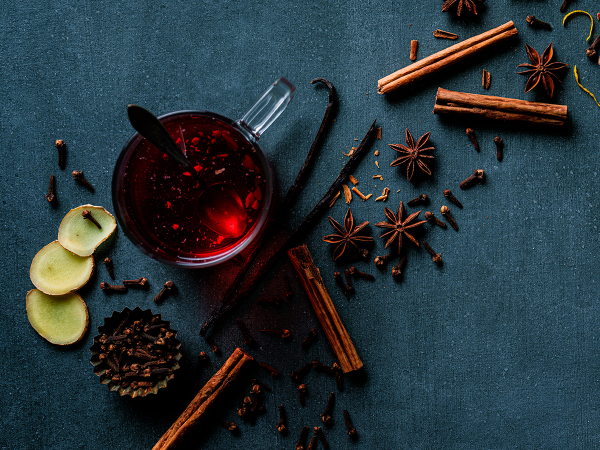 Kopp med varm gløgg omgitt av kanelstenger, stjerneanis, nellikspiker og epleskiver på en mørk bakgrunn.