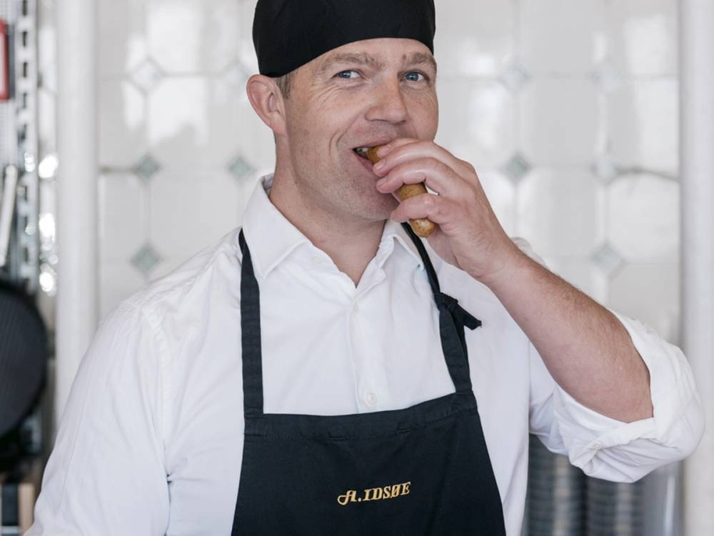 En person i hvit skjorte og svart forkle spiser en pølse, med en bakgrunn av et kjøkken eller matstasjon.