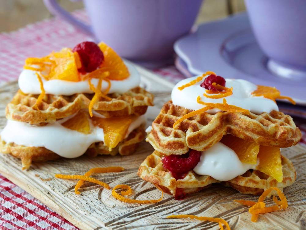 To glutenfrie vafler med krem, frukt og bær på en treplate, med kopper i bakgrunnen.