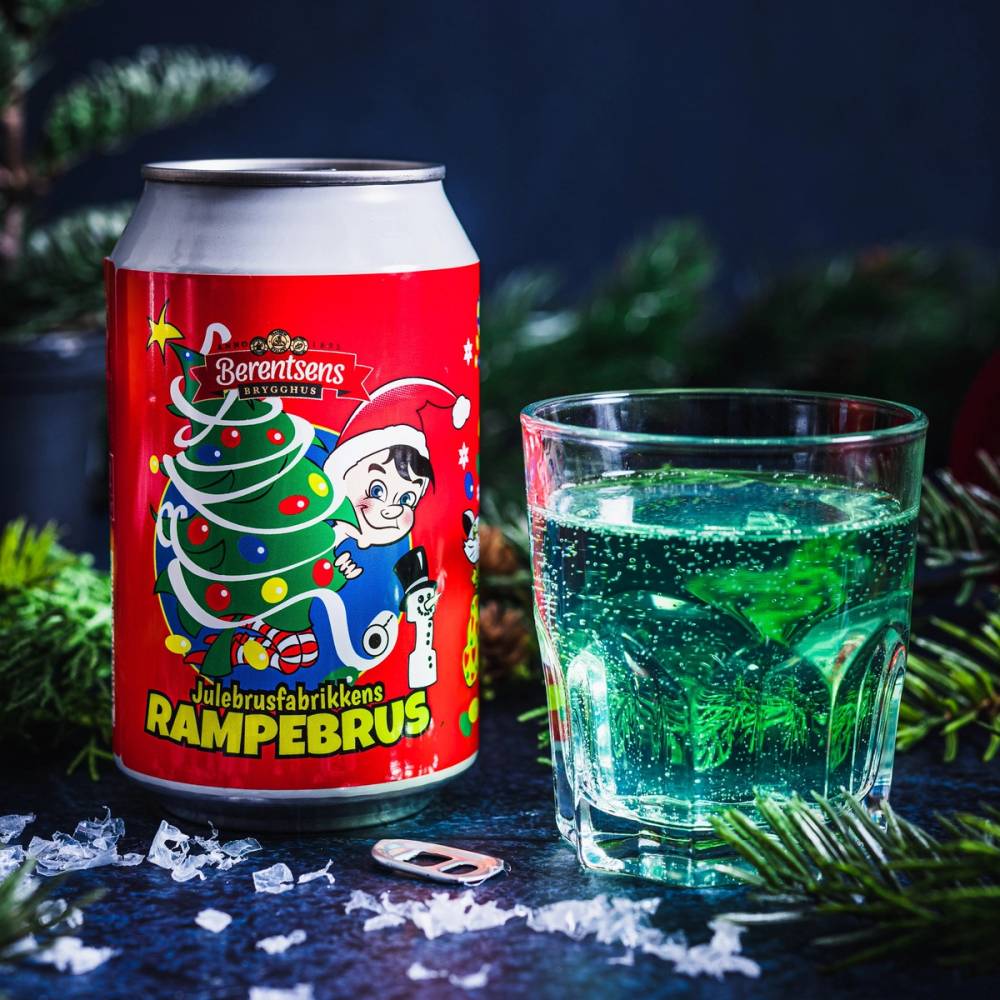 En rød julebrusboks med juledekorasjon og en glass med grønn brus, omgitt av granbar og snøaktige elementer.