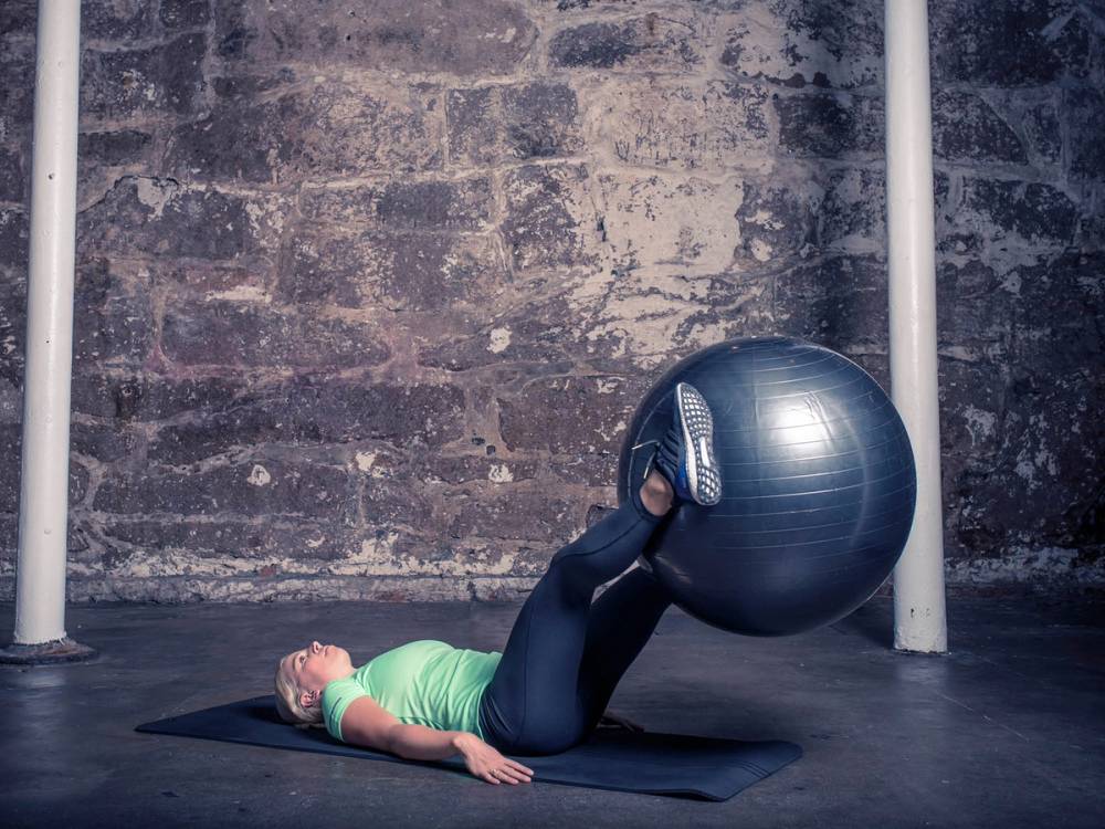 En person ligger på ryggen og løfter en stor treningsball med føttene i et treningsrom med murvegg.