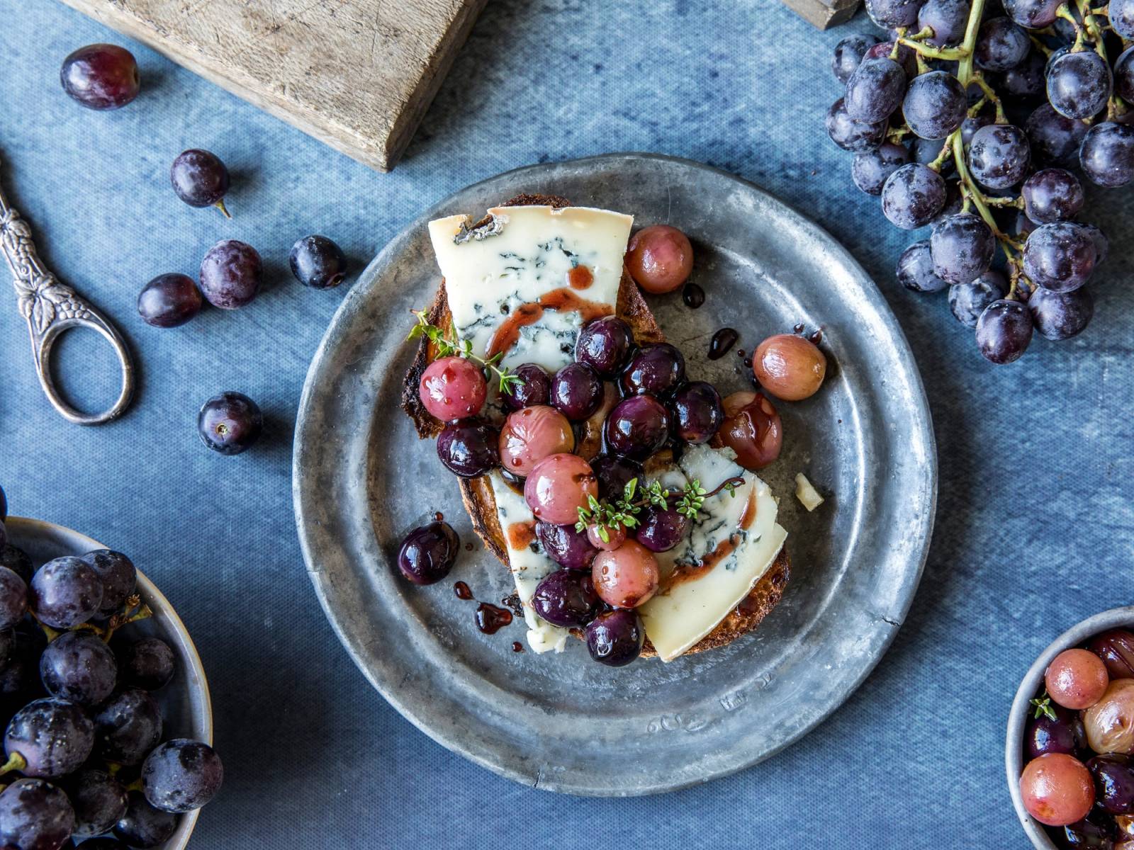 Ulike druer på en tallerken med ost, omgitt av flere druer og en saks på en blå bakgrunn.