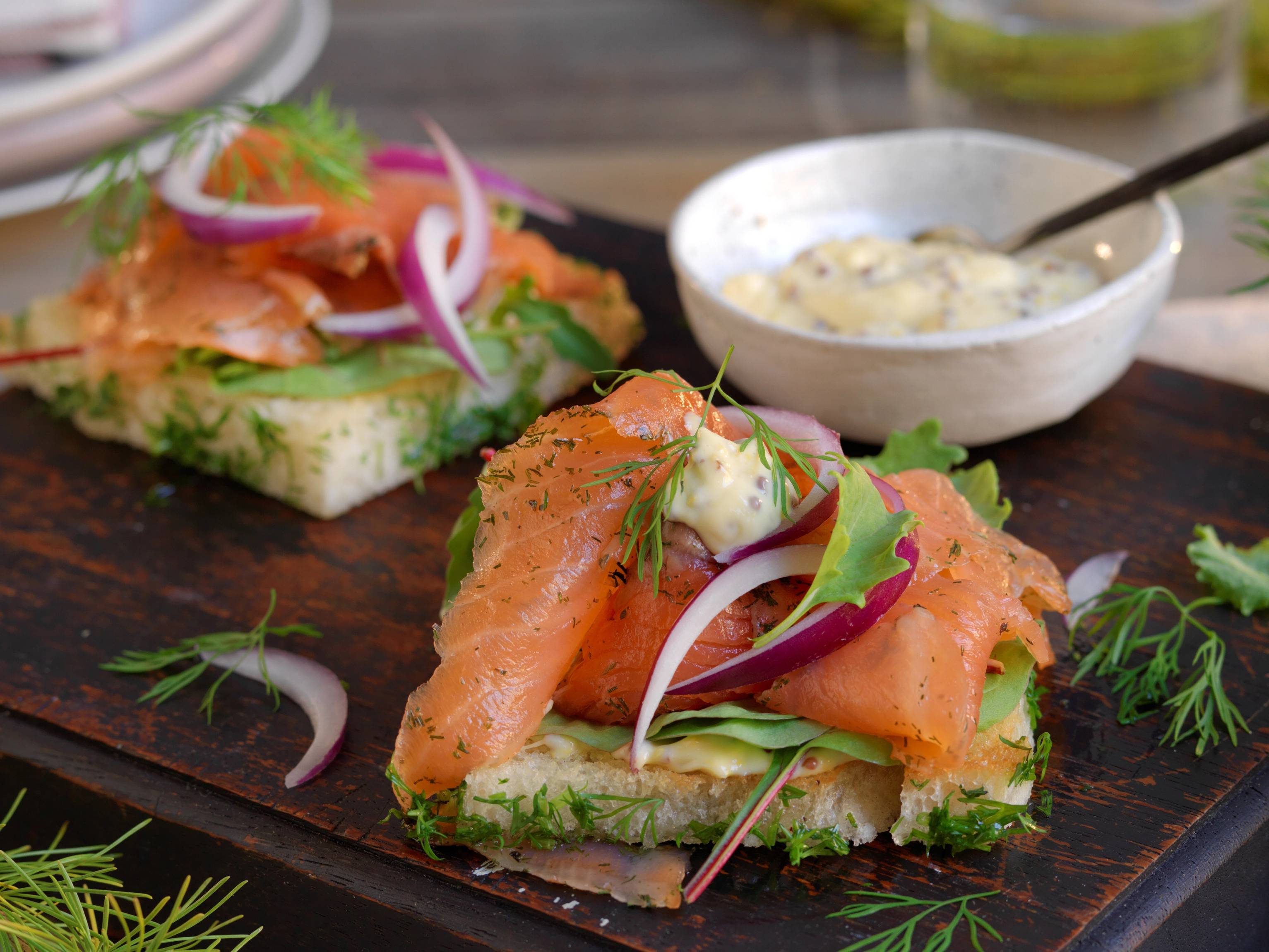 To åpne toast med gravlaks, kremost, rødløk og dill på en skjærebrett med en liten skål med saus ved siden av.
