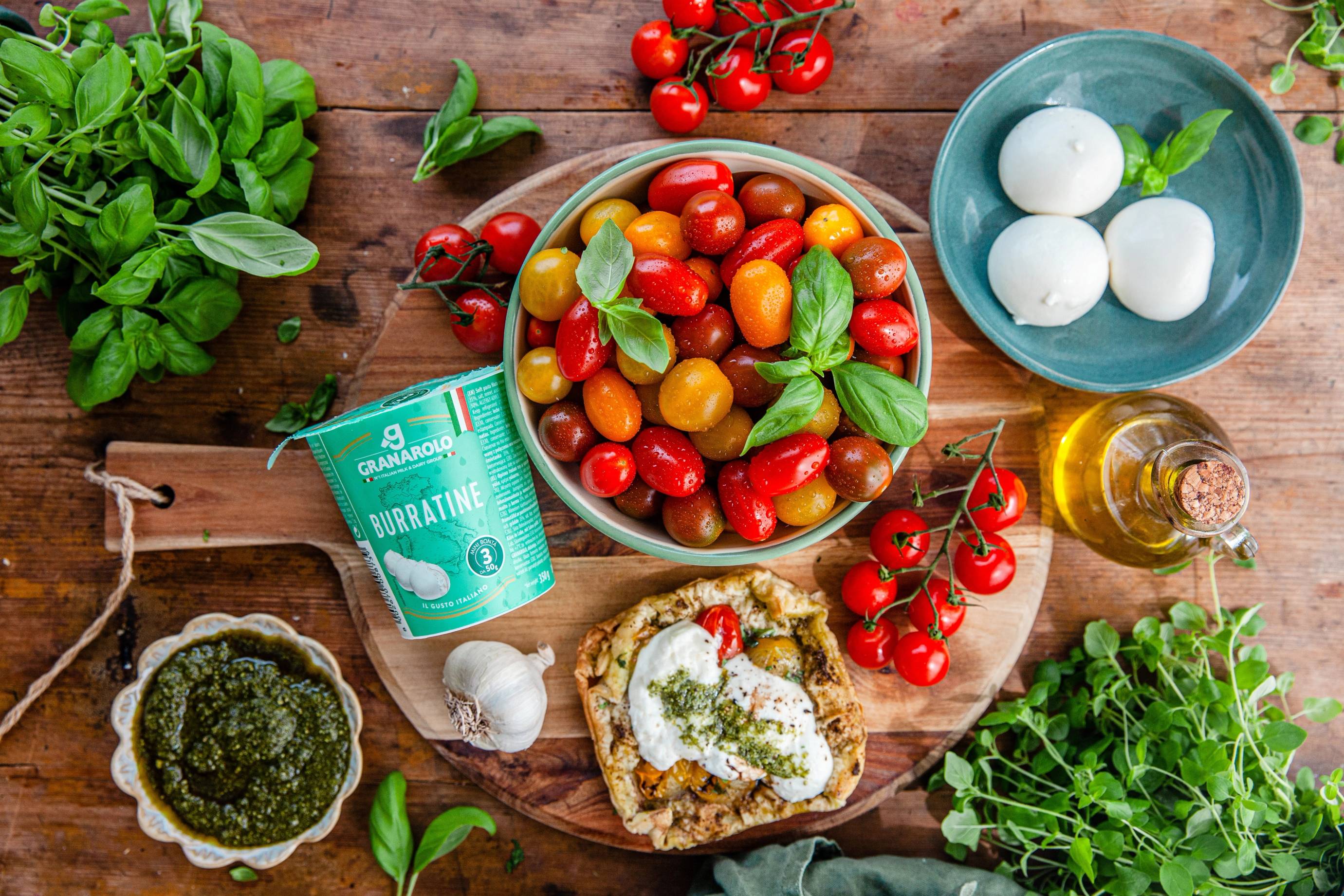 Et fat med ulike fargede cherrytomater, frisk basilikum, en skål med mozzarella, og brød med ost og urter på et trebord, omgitt av urter og tomater.