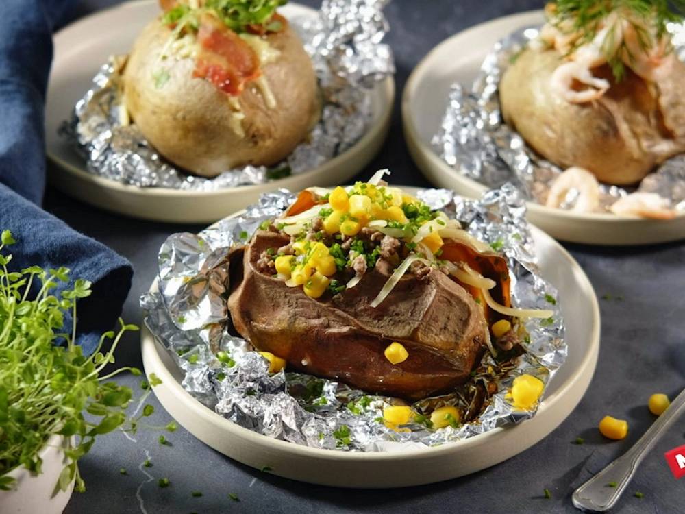 Bakte poteter med fyll, servert på små tallerkener med aluminiumsfolie, omgitt av grønne urter og tilbehør.