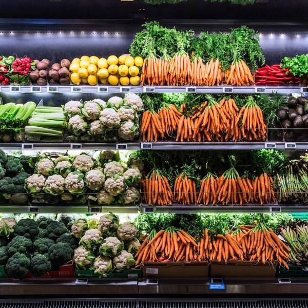 Et utvalg av ferske grønnsaker i en butikk, inkludert gulrøtter, brokkoli, blomkål, agurk og salat, arrangert på flere hyller.