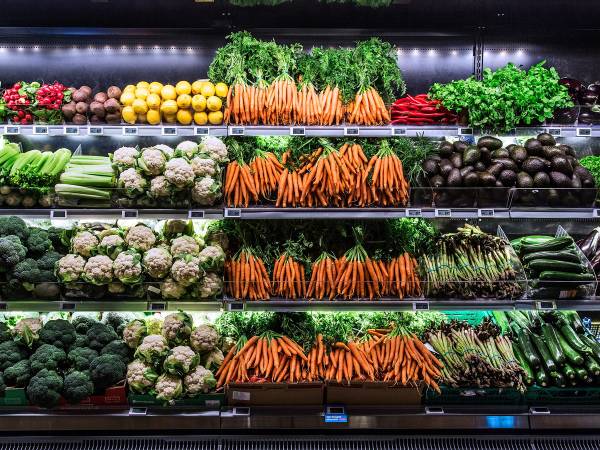 Et utvalg av ferske grønnsaker i en butikk, inkludert gulrøtter, brokkoli, blomkål, agurk og salat, arrangert på flere hyller.