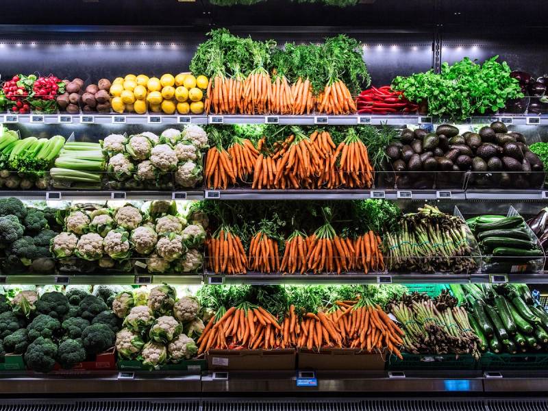 Et utvalg av ferske grønnsaker i en butikk, inkludert gulrøtter, brokkoli, blomkål, agurk og salat, arrangert på flere hyller.