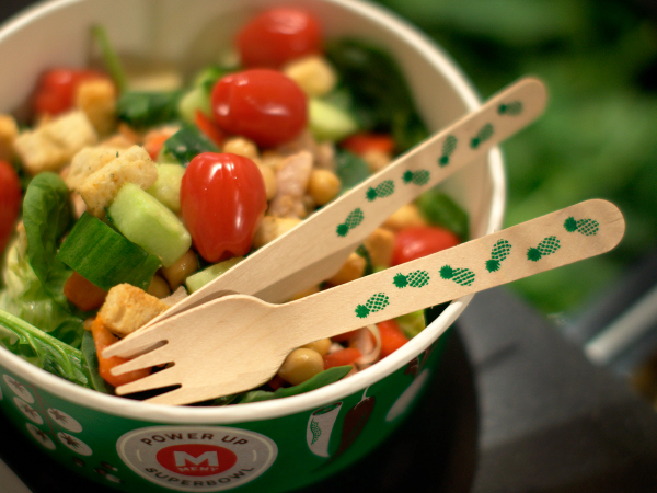 En salat i en grønn bolle med små tomater, mais og grønnsaker, med to engangsplastbestikk ved siden av.