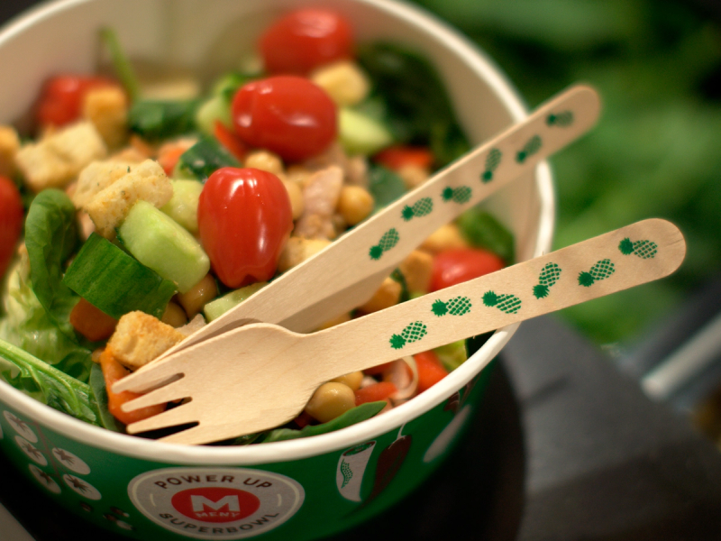 En salat i en grønn bolle med små tomater, mais og grønnsaker, med to engangsplastbestikk ved siden av.