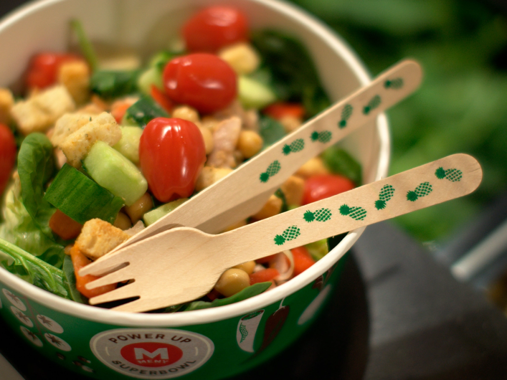 En salat i en grønn bolle med små tomater, mais og grønnsaker, med to engangsplastbestikk ved siden av.