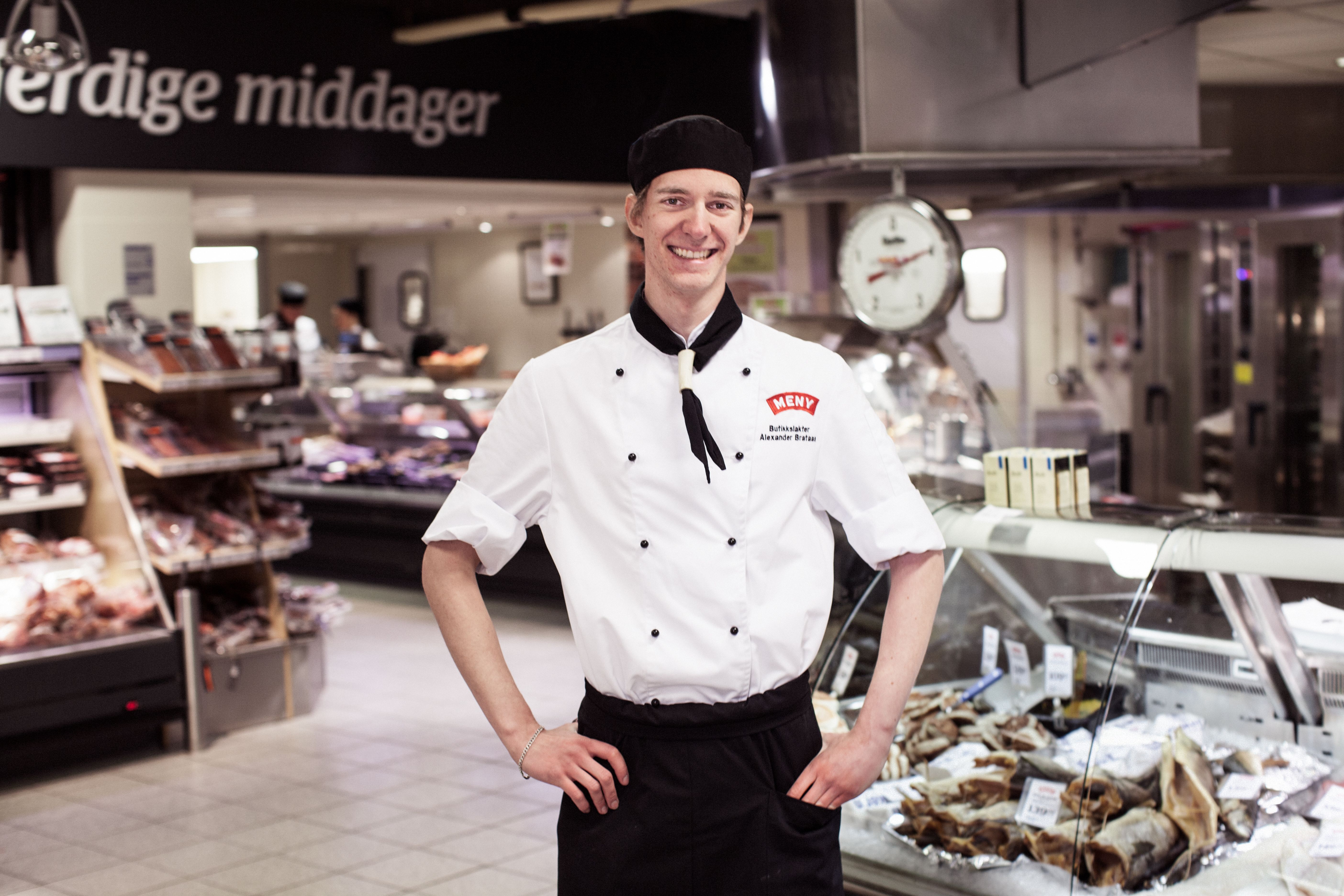 En person i kokkeforkle står i en matbutikk eller delikatesse med kjøtt og pålegg i bakgrunnen.