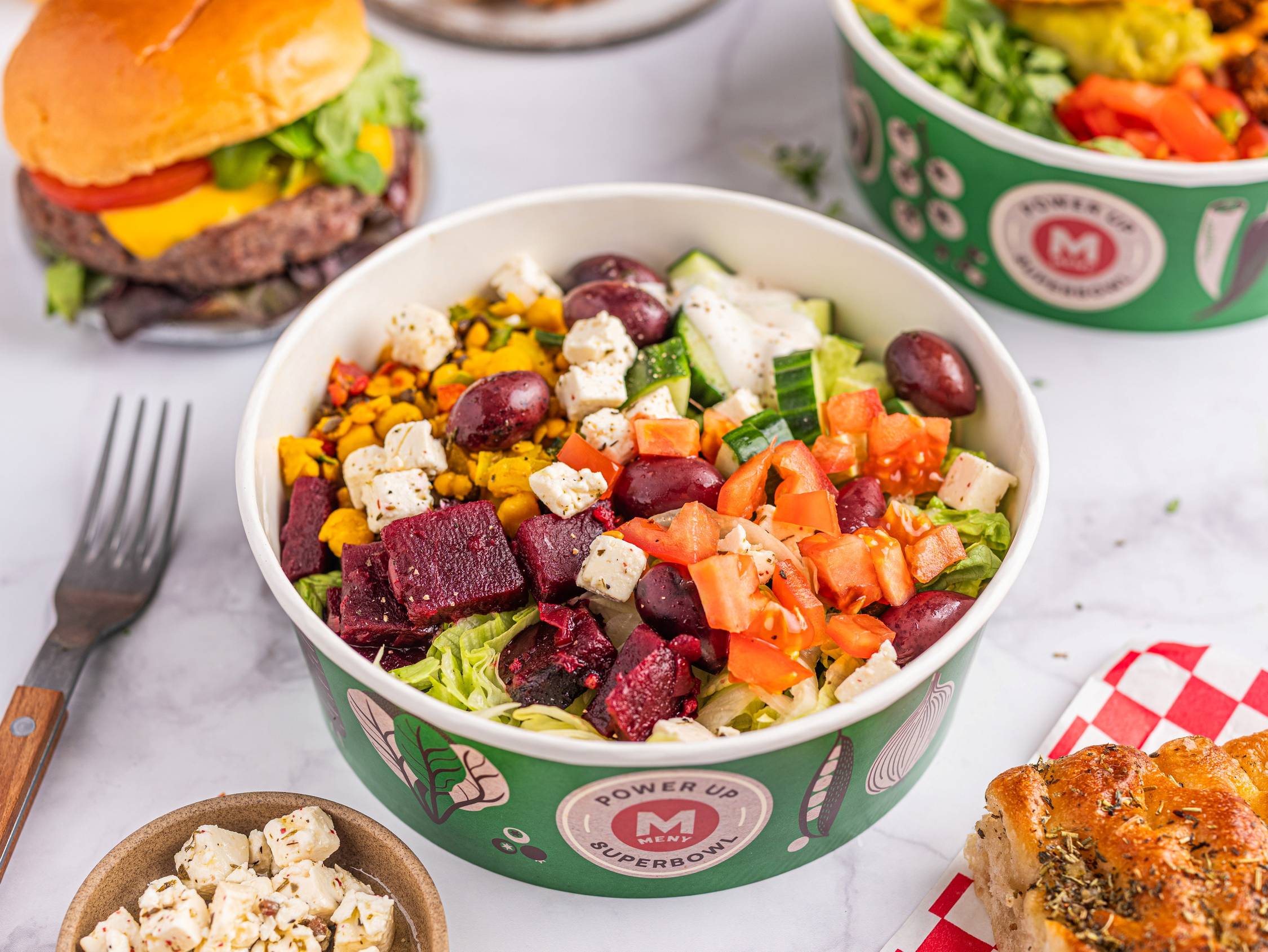 En salat med ulike grønnsaker og biter av ost i en grønn salatbolle, med en burger og en annen salat i bakgrunnen.