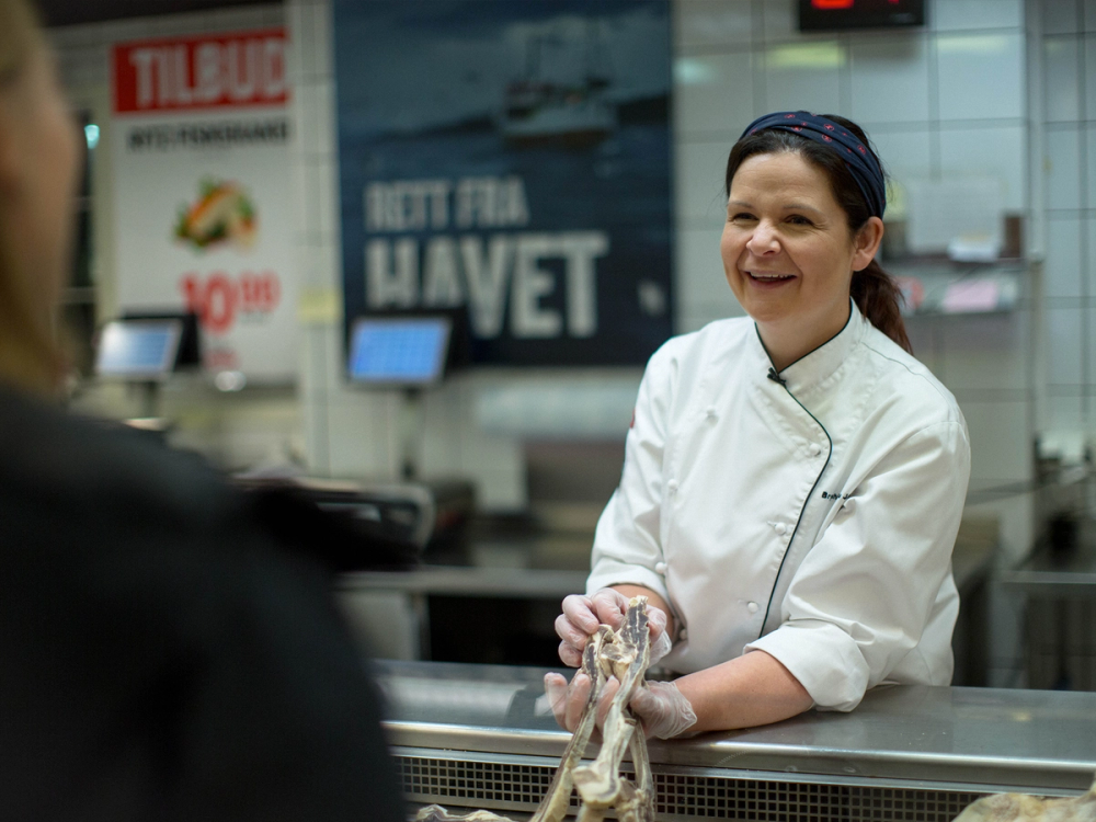 En person i kokkeforkle som håndterer sjømat i en butikk eller restaurant, med en plakat som sier 'Høtt fra havet' i bakgrunnen.