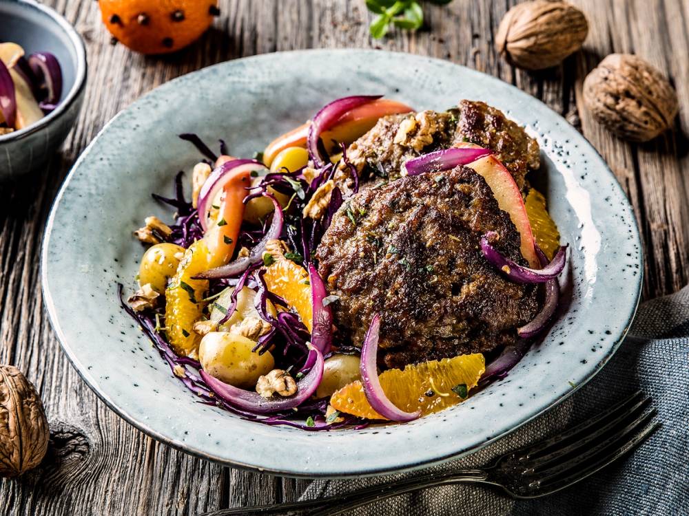 Karbonader med råkostsalat av gulrot og rødløk på en tallerken, omgitt av nøtter og små skåler med tilbehør.