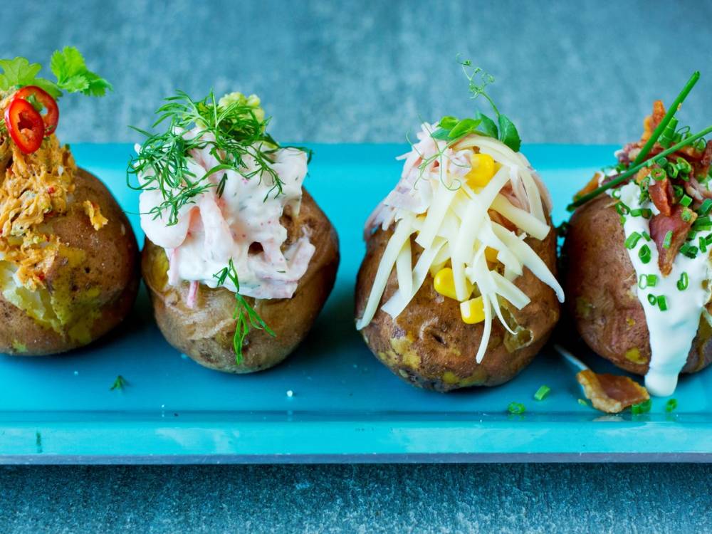 Fire bakte poteter med ulike toppings som ost, urter og grønnsaker på en blå serveringsfat.