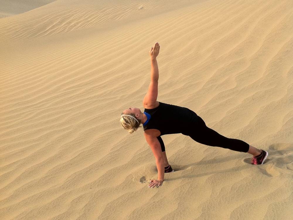 En person gjør en yoga- eller treningsøvelse på sanddyner, med en arm løftet opp og kroppen bøyd til siden.