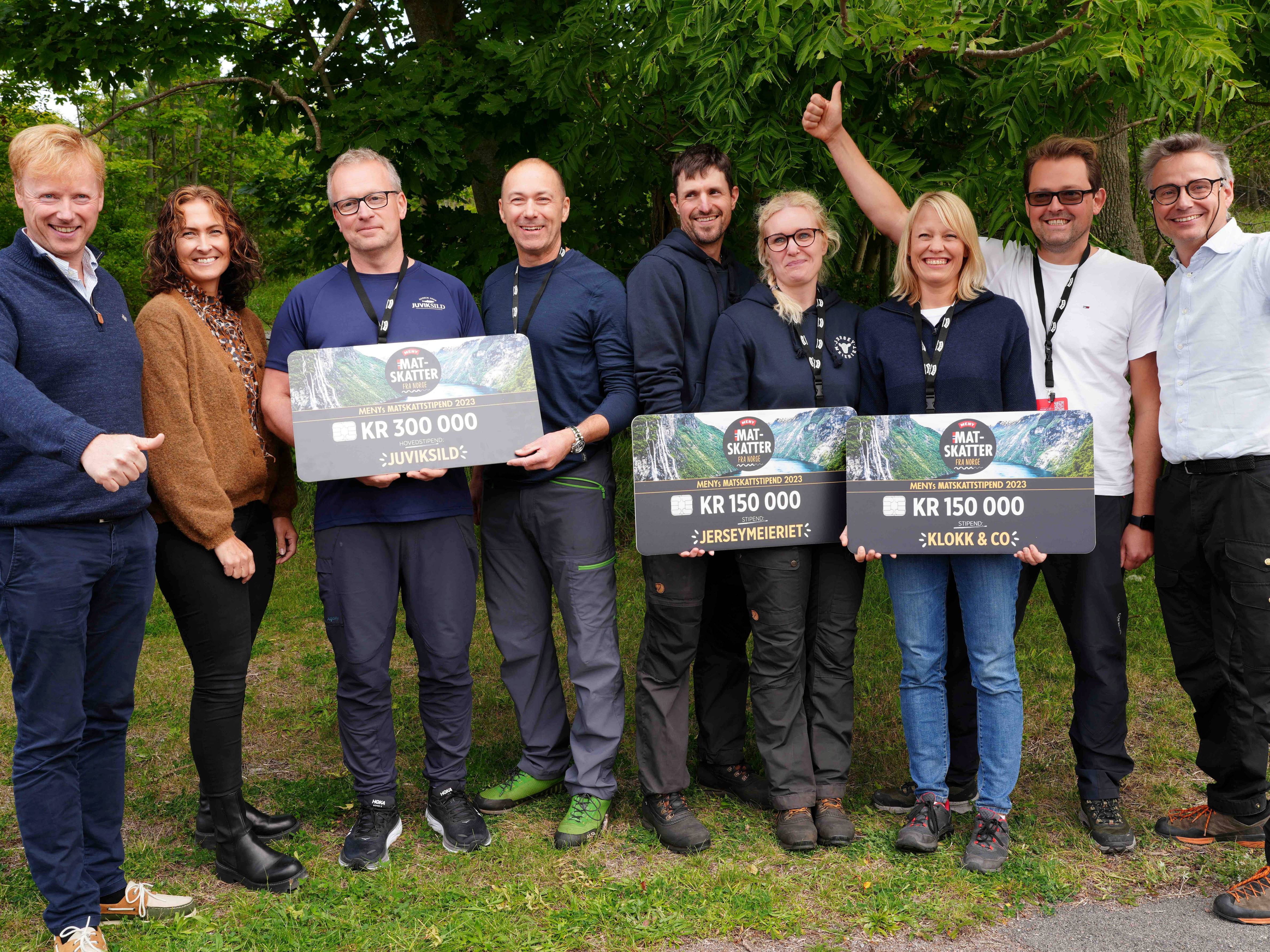 Gruppe med vinnere av Matskattstipendiet ved MENY 2023 som holder store sjekkbeløp, utendørs med grønne trær i bakgrunnen.
