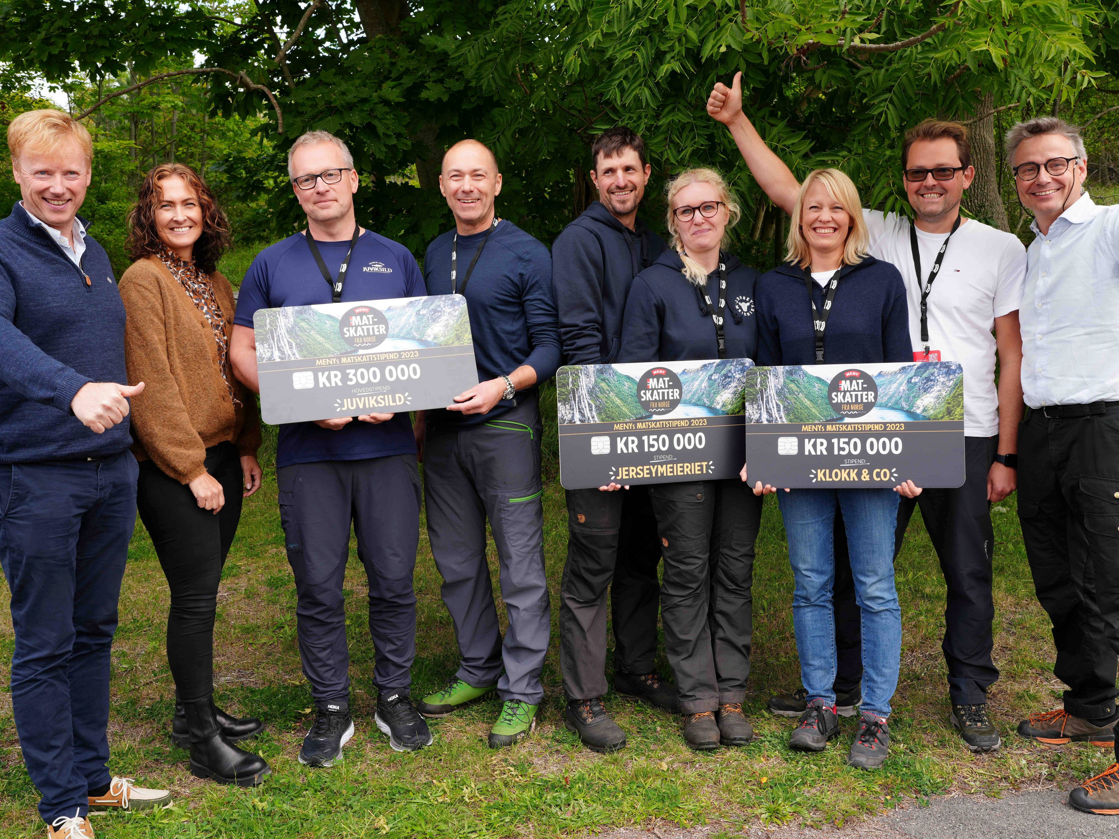 Gruppe med vinnere av Matskattstipendiet ved MENY 2023 som holder store sjekkbeløp, utendørs med grønne trær i bakgrunnen.