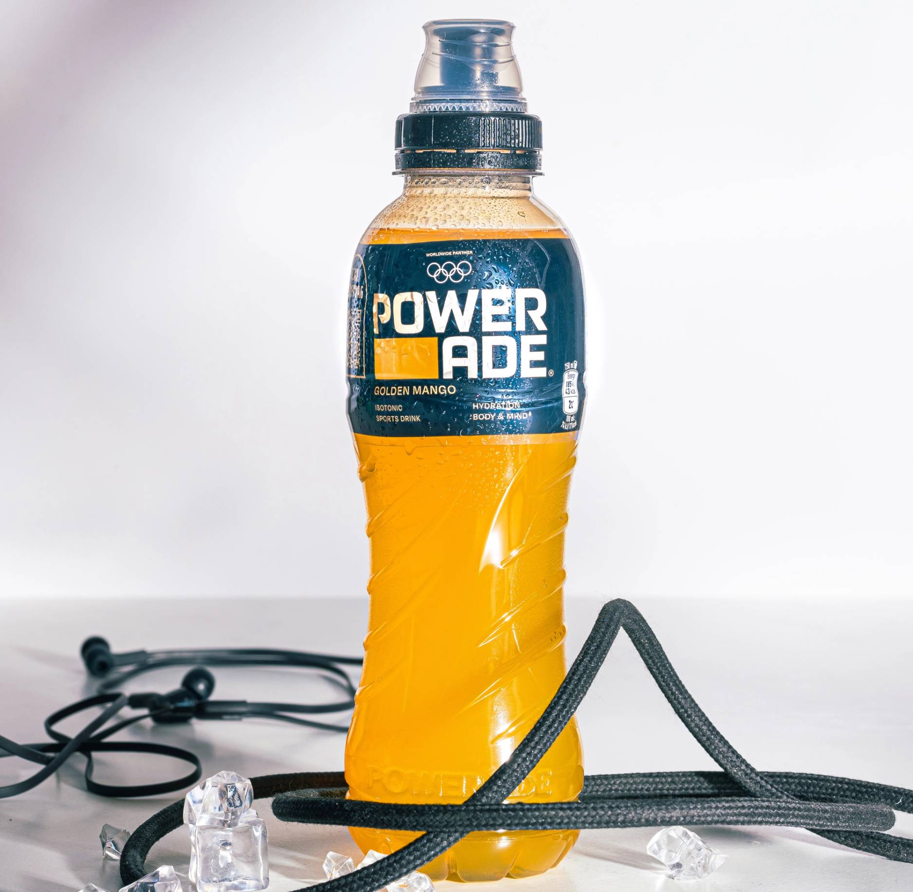 En flaske med Powerade mango-smak står på et bord, omgitt av hodetelefoner og treningsutstyr.