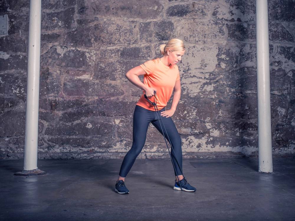 En kvinne utfører enarms roing med manual i et treningsstudio med betongvegg i bakgrunnen.