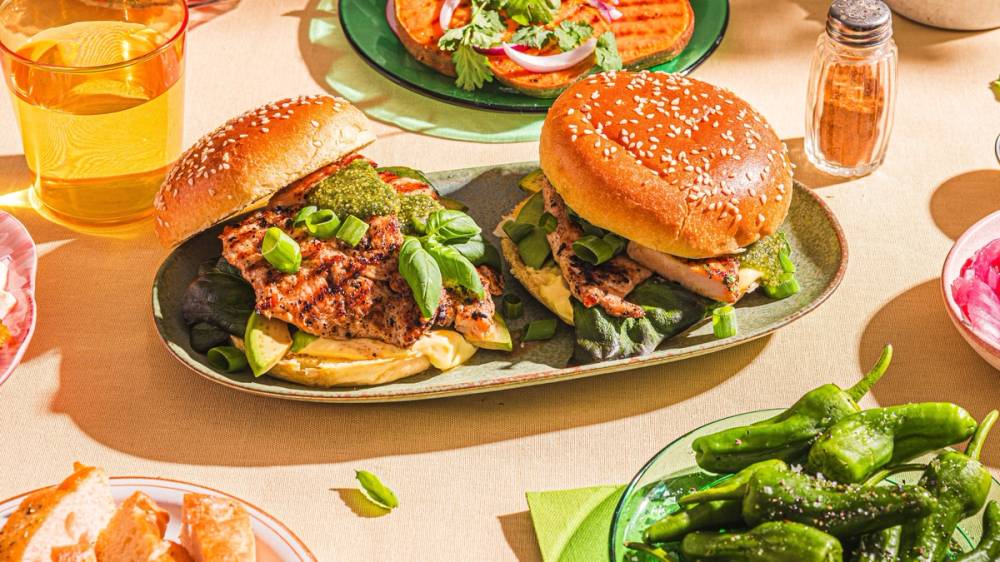 To burgere med salat og kjøttdeig på en tallerken, omgitt av grønnsaker og drikke, på et bord med sommerrammeplakat.