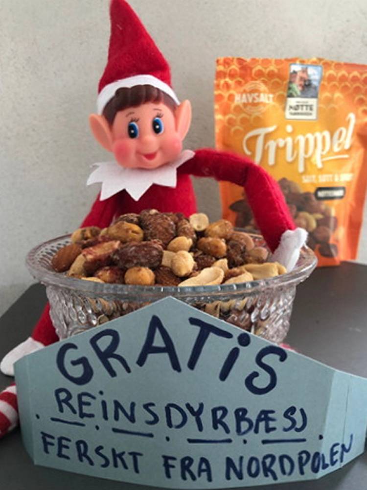 En liten nissefigur med rød hatt og klær foran en skål med nøtter, med en gratis reinsdyrbæsj-kort og en pose med trippel sjokolade i bakgrunnen.