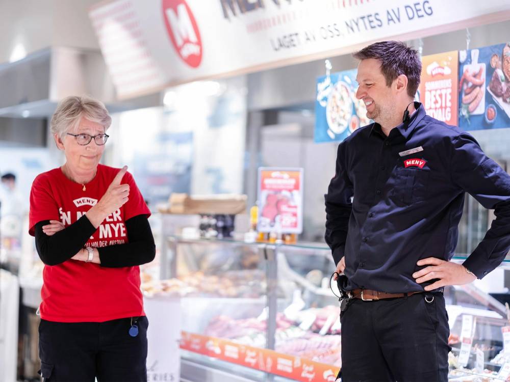 To personer snakker i en butikk, en av dem har på seg en svart jakke og den andre en rød genser med tekst, i en matbutikk med reklameskilt og matvarer i bakgrunnen.