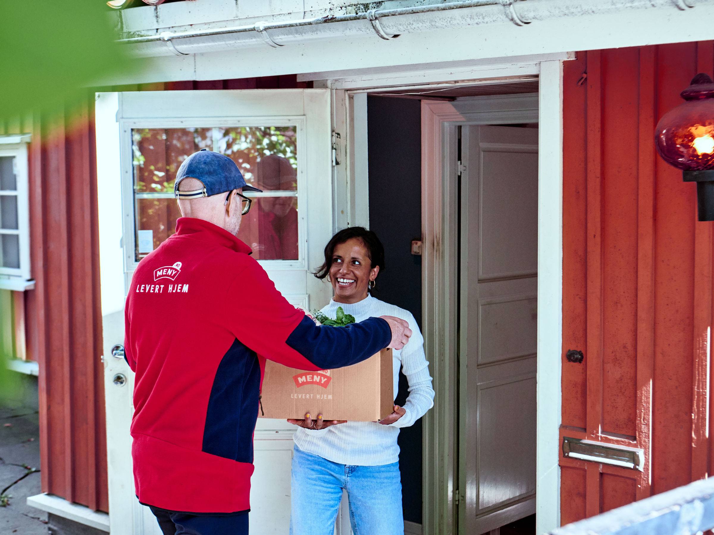 En person i rød jakke gir en pakke til en ung person utenfor en rød husvegg, med en åpen dør i bakgrunnen.